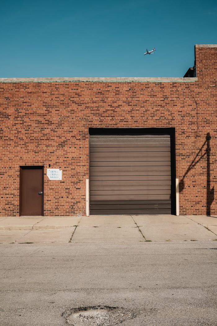 A brick industrial building with a garage door and airplane in the clear sky above.